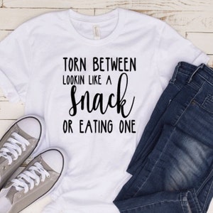 May include: White t-shirt with black text that reads "Torn between lookin like a snack or eating one".