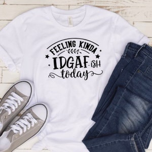 May include: White t-shirt with black text that reads "Feeling Kinda IDGAF-ish today". The shirt is laid out on a white wooden surface with a pair of gray sneakers and blue jeans.