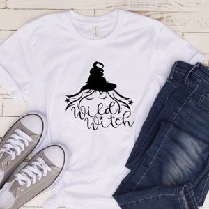 May include: White t-shirt with a black graphic of a witch with the text "Wild Witch" on it. The t-shirt is laying on a wooden surface with a pair of gray sneakers and blue jeans.