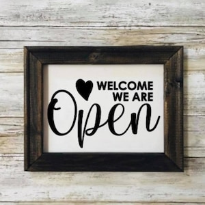 May include: A framed sign with a white background and black text that reads "WELCOME WE ARE Open" with a heart symbol. The frame is dark brown, and the background is a light wood.