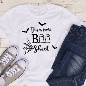 May include: White t-shirt with the text "this is some BOO Sheet" in black, along with bat and spiderweb graphics. The shirt is paired with blue jeans and gray sneakers, creating a casual Halloween-themed outfit.