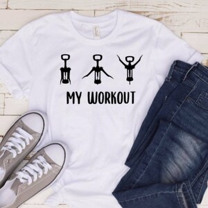 May include: White t-shirt with a graphic of three corkscrews and the text "MY WORKOUT". The t-shirt is laid out on a white wooden surface with a pair of grey sneakers and blue jeans.