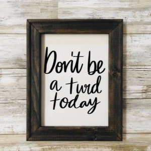 May include: A framed sign with the text "Don't be a turd today" in black cursive lettering on a white background. The frame is dark brown wood.