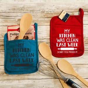 May include: Two red and blue kitchen pot holders with the text "My kitchen was clean last week. Sorry you missed it!"  The pot holders are on a wooden surface with kitchen utensils.