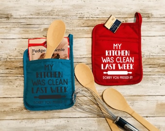 May include: Two red and blue kitchen pot holders with the text "My kitchen was clean last week. Sorry you missed it!"  The pot holders are on a wooden surface with kitchen utensils.