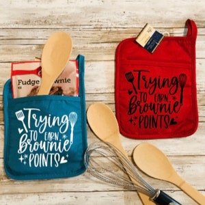May include: Two oven mitts, one teal and one red, with the text "Trying to Earn Brownie Points." Each mitt has a pocket containing a wooden spoon and brownie mix. Additional wooden spoons and a whisk are also present.