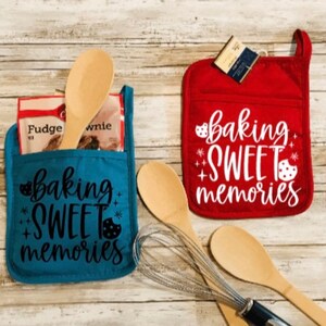 May include: Two blue and red oven mitts with the text "baking sweet memories" on them. The mitts are on a wooden surface with wooden spoons and a whisk.