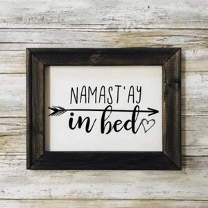 May include: A framed sign with a black arrow and a heart, with the text "Namast'ay in bed".
