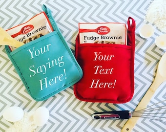 May include: Two red and blue oven mitts with pockets for holding a box of brownie mix. The mitts are personalized with the text "Your Saying Here!" and "Your Text Here!"