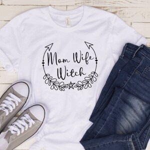May include: White t-shirt with black text that reads "Mom Wife Witch" in a circle with arrows and floral accents.