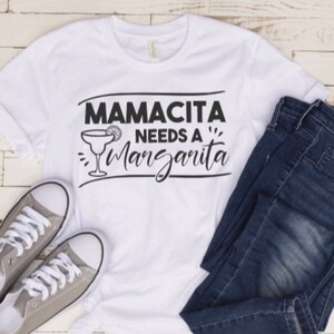 May include: White t-shirt with black text that reads "Mamacita Needs a Margarita" and a graphic of a margarita glass with a lime wedge. The shirt is laid out on a white wooden surface with a pair of gray canvas shoes and blue jeans.