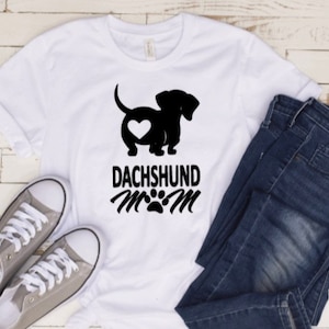 May include: White t-shirt with a black silhouette of a dachshund with a heart on its rear end. The text "Dachshund Mom" is printed below the dog with a paw print.