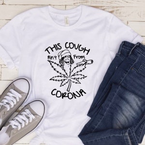 May include: White t-shirt with a black graphic of a cartoon person smoking a joint with the text "This Cough Ain't From Corona".
