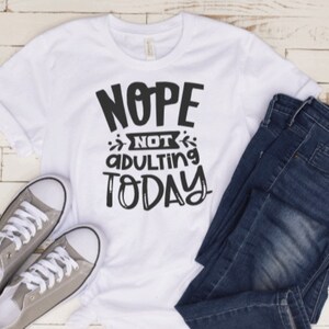 May include: White t-shirt with black text that reads "NOPE NOT ADULTING TODAY". The shirt is laying on a white wooden surface with a pair of gray canvas shoes and blue jeans.