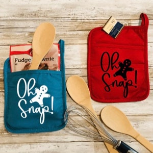 May include: Two red and blue oven mitts with a gingerbread man design and the text "Oh Snap!" printed on them. The mitts are on a wooden surface with wooden spoons and a whisk.