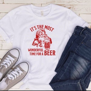May include: White t-shirt with a red graphic of Santa Claus holding a beer mug. The text reads "It's the most wonderful time for a beer".