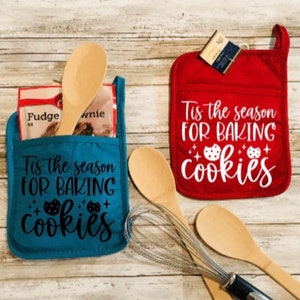 May include: Two blue and red oven mitts with a pocket on the front. The mitts have the text "Tis the season for baking cookies" printed on them. The mitts are on a wooden surface with wooden spoons and a whisk.