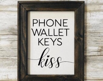 May include: A framed sign with the text "PHONE WALLET KEYS kiss" in black lettering on a white background. The frame is dark brown wood.