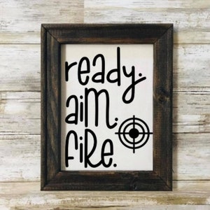 May include: A rustic wooden framed sign with the text "ready aim fire."  A black target symbol is next to the text.