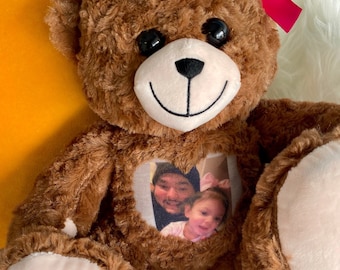 Può includere: Un orsacchiotto di peluche marrone con un fiocco rosa è seduto su una superficie bianca e soffice. L'orsacchiotto ha una finestra a forma di cuore sul petto con una foto di due persone all'interno.