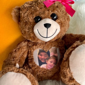 Puede incluir: Un oso de peluche marrón con un lazo rosa está sentado sobre una superficie blanca y esponjosa. El oso tiene una ventana en forma de corazón en su pecho con una foto de dos personas dentro.