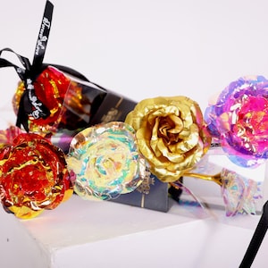 May include: A collection of colorful, iridescent artificial roses. The roses are in shades of red, gold, and purple. Each rose has a metallic sheen and is presented with a black ribbon that reads "Forever Love".