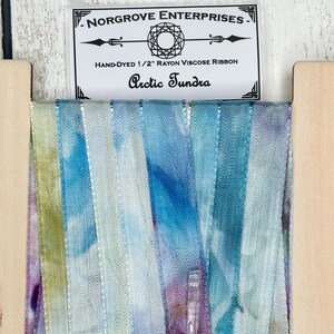 May include: A close-up of a spool of hand-dyed rayon viscose ribbon in shades of blue, green, and purple. The ribbon is labeled "Arctic Tundra" and is from Norgrove Enterprises.