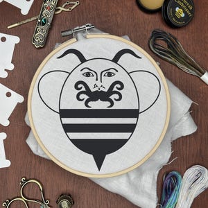 May include: An embroidery hoop with a black and white bee design, surrounded by embroidery supplies. Includes scissors, thread, a thread conditioner, and a needle case. The bee design has a face and mustache.