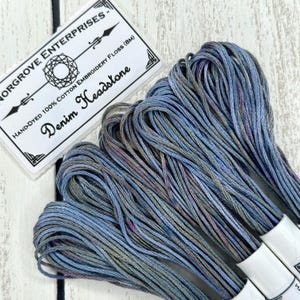 Könnte beinhalten: Nahaufnahme von handgefärbtem Baumwoll-Stickgarn in Blau-, Grau- und Lilatönen. Das Etikett trägt die Aufschrift "Denim Headstone" und "Hand-Dyed 100% Cotton Embroidery Floss (8M)". Das Garn ist gebündelt und bereit für Stickprojekte.