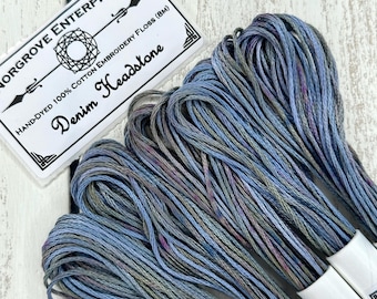 May include: Close-up of hand-dyed cotton embroidery floss in shades of blue, gray, and purple. The label reads "Denim Headstone" and "Hand-Dyed 100% Cotton Embroidery Floss (8M)". The floss is bundled and ready for use in embroidery projects.