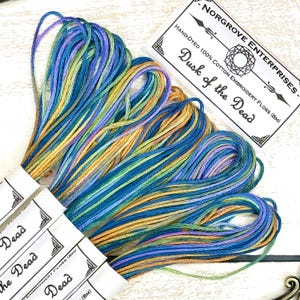 以下が含まれることがあります： 青、緑、金色の手染め刺繍糸の束。「Dusk of the Dead」とラベルされており、綿100％です。画像には、ラベルが見える糸の束がいくつか表示されています。