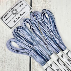 May include: A close-up of several spools of hand-dyed cotton embroidery floss in shades of blue and purple. The spools are labeled "The Polite Doctrine" and "Norgrove Enterprises".
