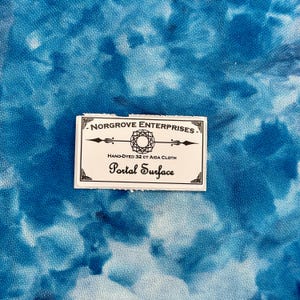 May include: A piece of hand-dyed 32 ct Aida cloth featuring a mottled blue and white pattern. A rectangular white label is centered on the fabric with the text Norgrove Enterprises, Hand-Dyed 32 ct Aida Cloth, and Portal Surface printed.