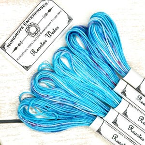 Könnte beinhalten: Mehrere Stränge handgefärbten Baumwoll-Stickgarns in Blau- und Weißtönen mit violetten Tupfen. Das Garn ist mit "Reactor Water" und "Norgrove Enterprises" beschriftet. Das Bild zeigt mehrere Stränge des 100% Baumwollgarns.