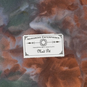 May include: A piece of hand-dyed 22 ct Aida cloth with a mottled orange, brown, and green pattern. A white rectangular label is centered on the fabric, reading Norgrove Enterprises, Hand-Dyed 22 ct Aida Cloth, and the color name Mud Pit.