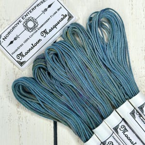 May include: Bundles of hand-dyed cotton embroidery floss in shades of blue, green, and purple. The floss is labeled "Moonstorm Masquerade" and "Norgrove Enterprises." The floss is 8 meters in length.