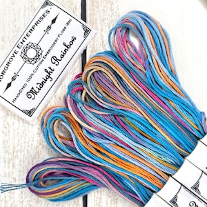 May include: A skein of hand-dyed 100% cotton embroidery floss in a variegated colorway of blue, orange, pink, and purple. The label reads "Midnight Rainbow" and "Hand-Dyed 100% Cotton Embroidery Floss (8M)" by "Orgrove Enterprises".