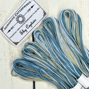 Könnte beinhalten: Handgefärbtes Stickgarn in Blau-, Weiß- und Beigetönen. Das Etikett trägt die Aufschrift "Sky Captain" und "NORGROVE ENTERPRISES". Auf dem Etikett steht außerdem, dass das Garn aus 100 % Baumwolle besteht und 8 Meter lang ist.