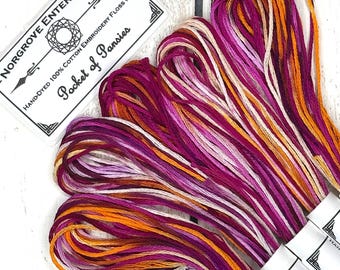 May include: A collection of hand-dyed 100% cotton embroidery floss in shades of magenta, purple, orange, and cream. The label reads Norgrove Enterprises, Hand-Dyed 100% Cotton Embroidery Floss (8M), and Pocket of Pansies. The threads are bundled.