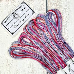 May include: A collection of hand-dyed cotton embroidery floss in red, pink, and blue hues. The label reads "Flame Forged Iron" from Norgrove Enterprises. The skeins are bundled, displaying the colour variations.
