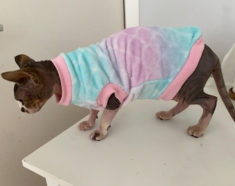 May include: A Sphynx cat wearing a tie-dye sweater in shades of blue, pink, and purple. The sweater has pink trim around the neck and armholes. The cat is standing on a white surface.
