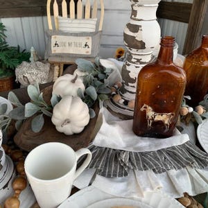 May include: A rustic farmhouse table setting with white pumpkins, a wooden bowl, a white mug, and two brown glass bottles with a horse design. The table is decorated with a white tablecloth with a gray ruffle and a garland of wooden beads.