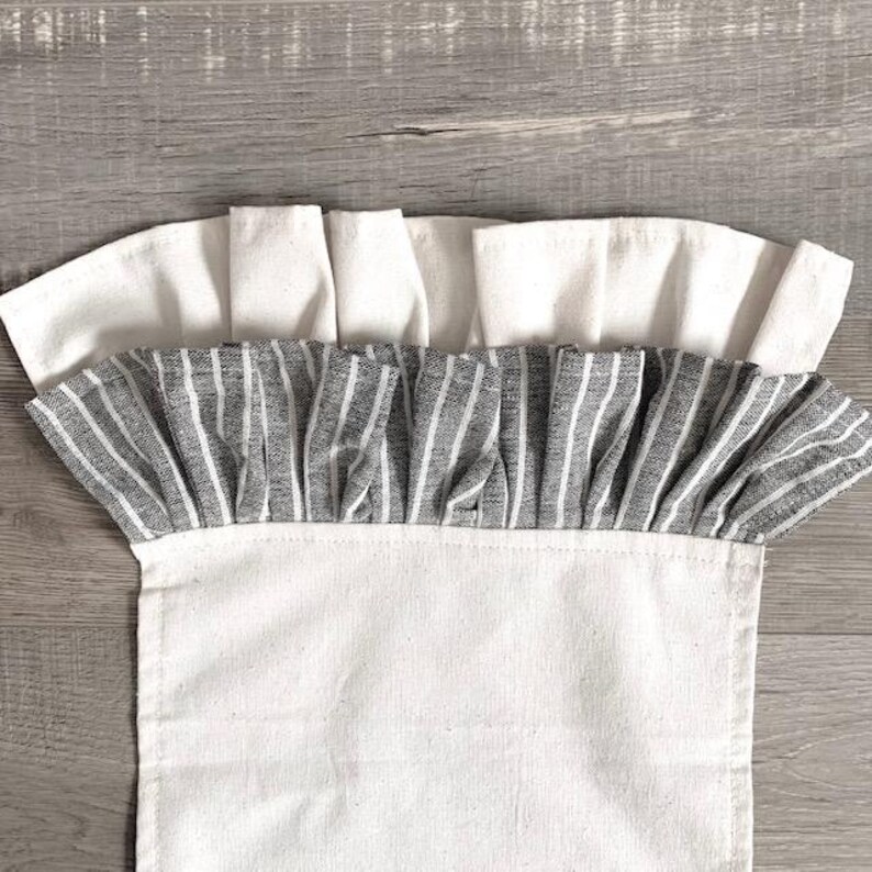 May include: A white fabric kitchen towel with a gray and white striped ruffle trim. The towel is folded and displayed on a wooden surface.