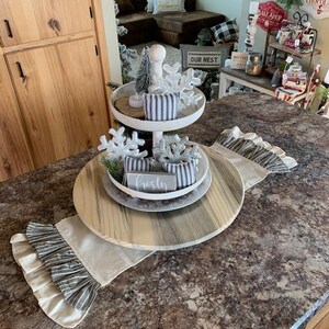 May include: A two-tiered white wooden serving tray with a wooden base. The tray is decorated with a small artificial pine tree, a white snowflake, and a white sign that says "Frosty". The tray is sitting on a table with a white and gray striped table runner.