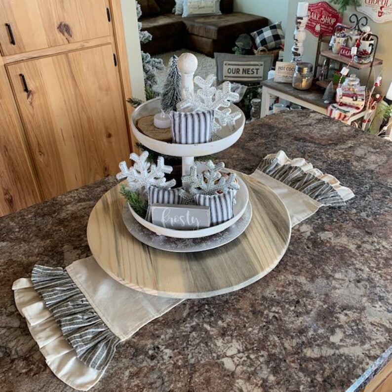 May include: A two-tiered white wooden serving tray with a wooden base. The tray is decorated with a small artificial pine tree, a white snowflake, and a white sign that says "Frosty". The tray is sitting on a table with a white and gray striped table runner.
