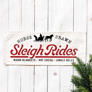 May include: A rectangular, off-white fabric sign with the words "HORSE DRAWN Sleigh Rides" in red and black lettering. The sign features a silhouette of a horse-drawn sleigh and the phrases "WARM BLANKETS - HOT COCOA - JINGLE BELLS".