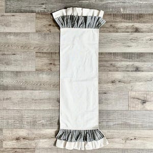 May include: White table runner with a black and white striped ruffle trim. The runner is made of a soft, lightweight fabric and is perfect for adding a touch of elegance to any table setting.