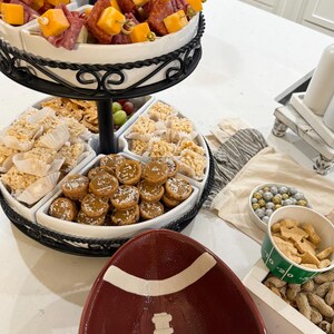May include: A three-tiered serving tray with a variety of snacks, including salami and cheese skewers, rice crispy treats, and small cookies. The tray is on a white countertop with a football-shaped bowl and a bowl of peanuts.
