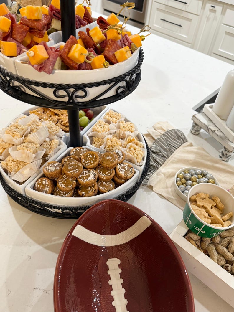May include: A three-tiered serving tray with a variety of snacks, including salami and cheese skewers, rice crispy treats, and small cookies. The tray is on a white countertop with a football-shaped bowl and a bowl of peanuts.