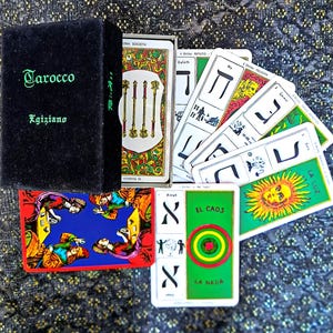 Può includere: Una scatola nera con l'etichetta Tarocco Egiziano contenente un mazzo di tarocchi. Le carte presentano illustrazioni colorate, lettere ebraiche e testo tra cui La Luz, El Caos e La Nada. Alcune carte sono disposte a ventaglio.